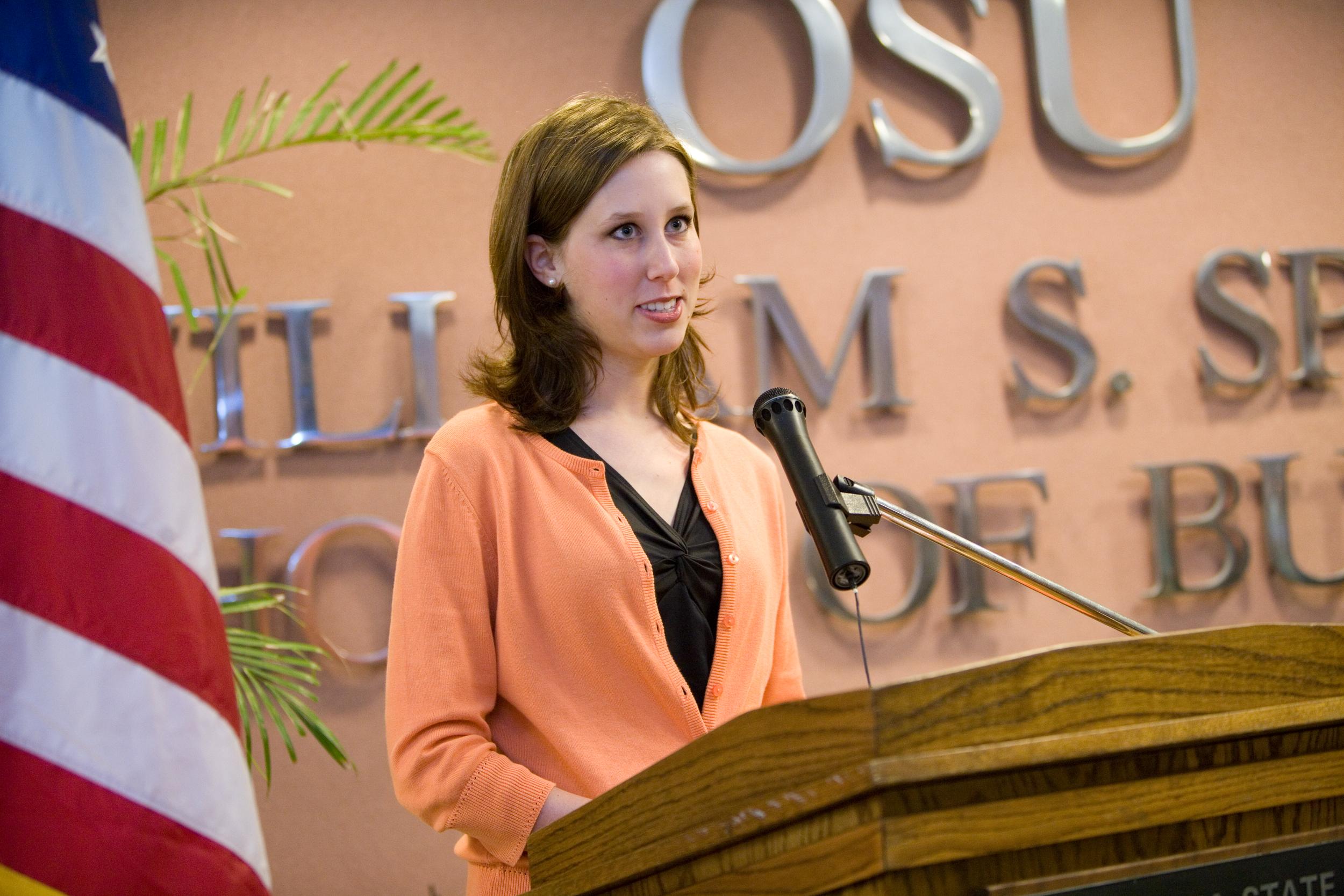 A young white woman stands at a lectern with a microphone at an event