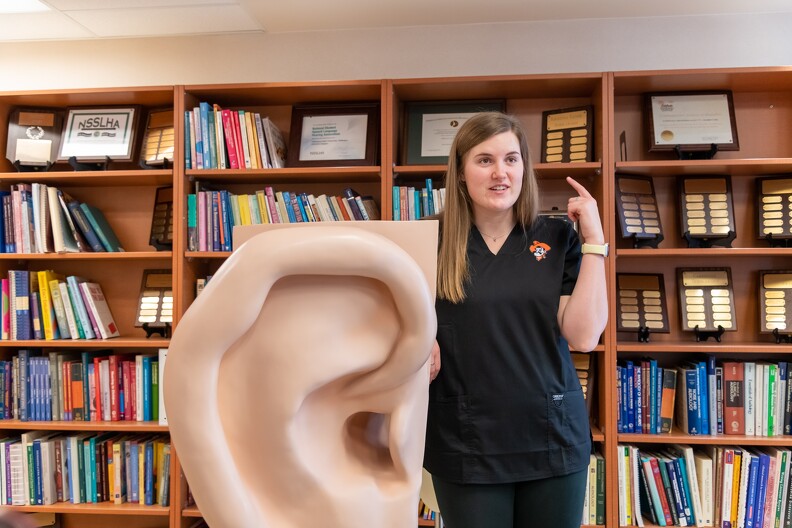 A female college student stands in front of a large model of the human ear as she gives an informative presentation.