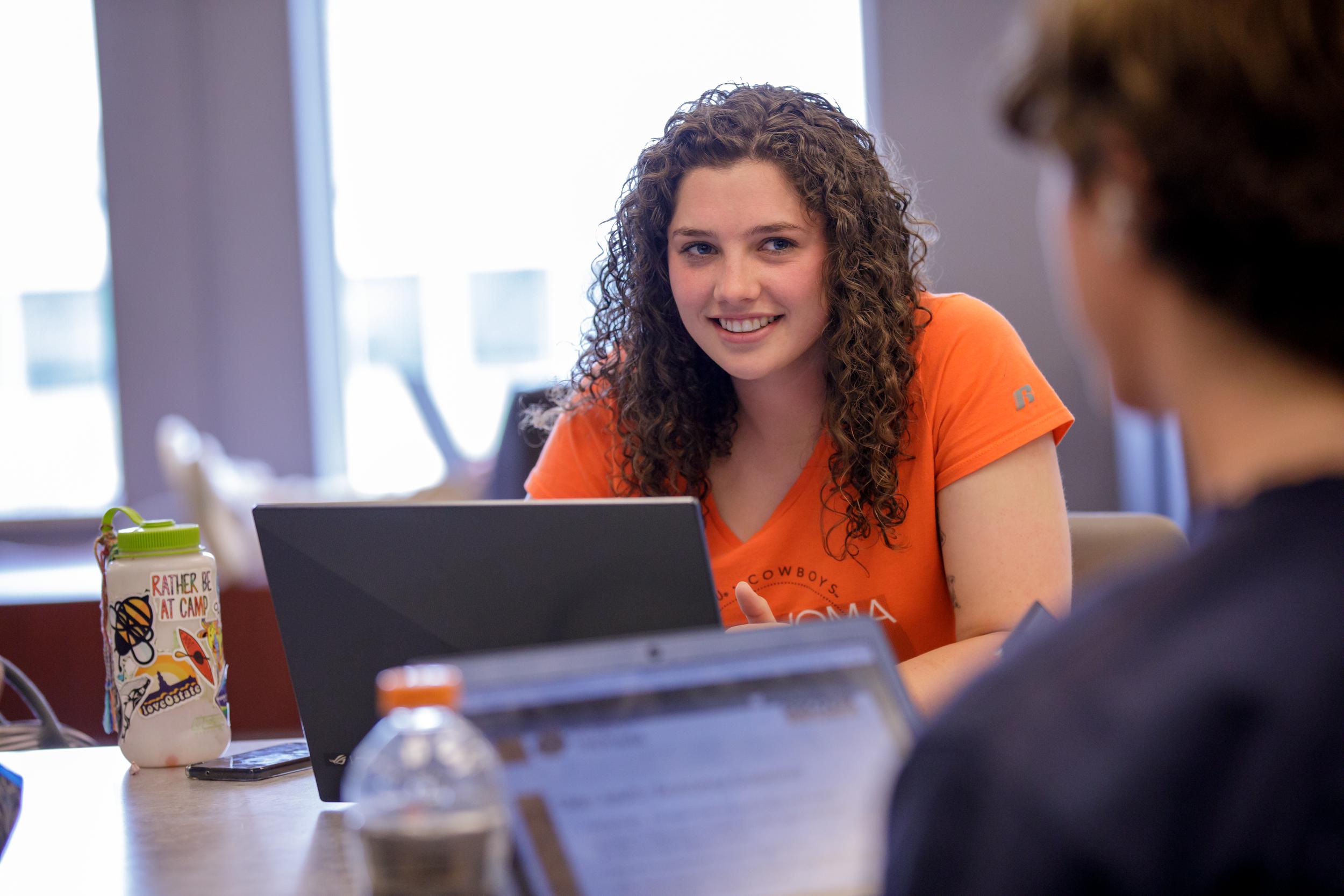 Young woman sitting at a table with a laptop in front of her while smiling at a person sitting on the other side of the table