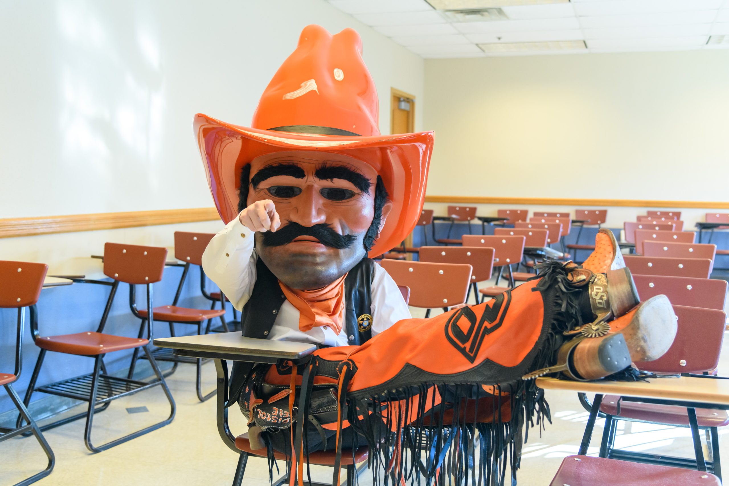 Pistol Pete is sitting with his legs crossed and on a student desk while pointing to the camera