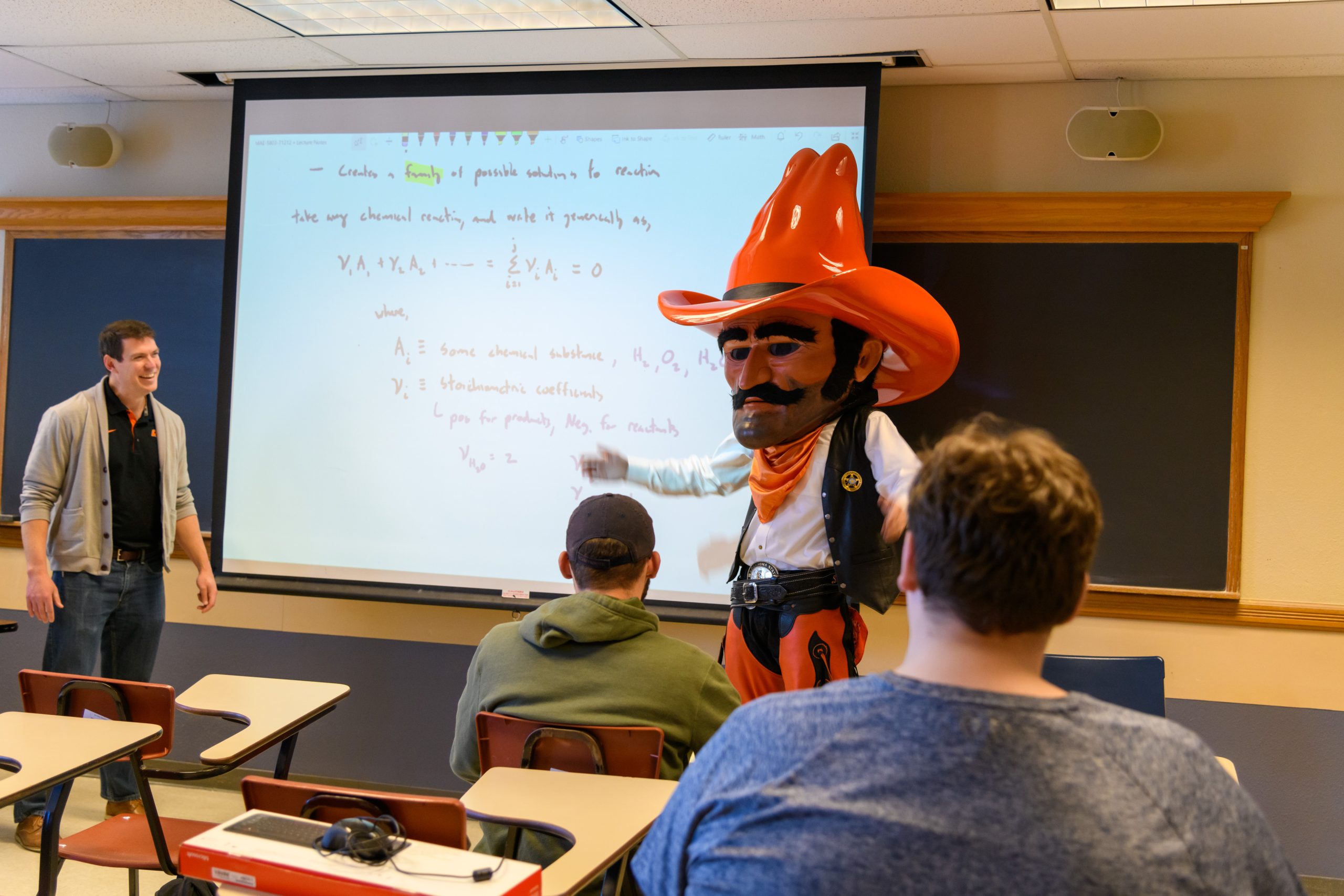 Pistol Pete stands at the front of the classroom pointing to a thermodynamics illustration on the projector screen to clarify difficult material.