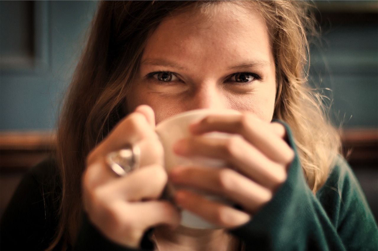 A woman with her hands wrapped around a coffee cup pulled to her mouth shows amusement or happiness with her eyes.