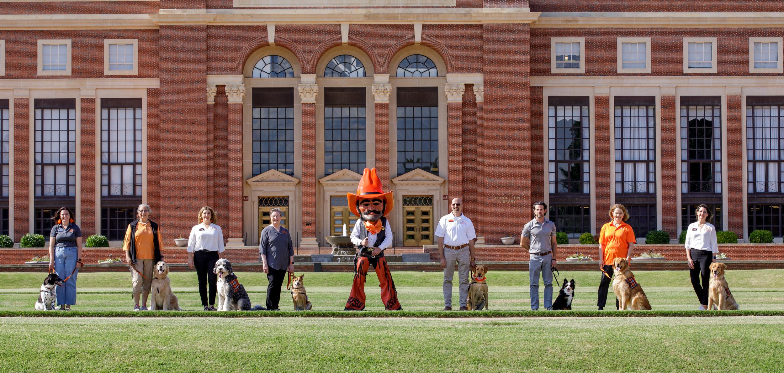 A group of Pete's Pet Posse dogs and their owners stand in a line with Pistol Pete on the library lawn in front of Edmon Low Library.