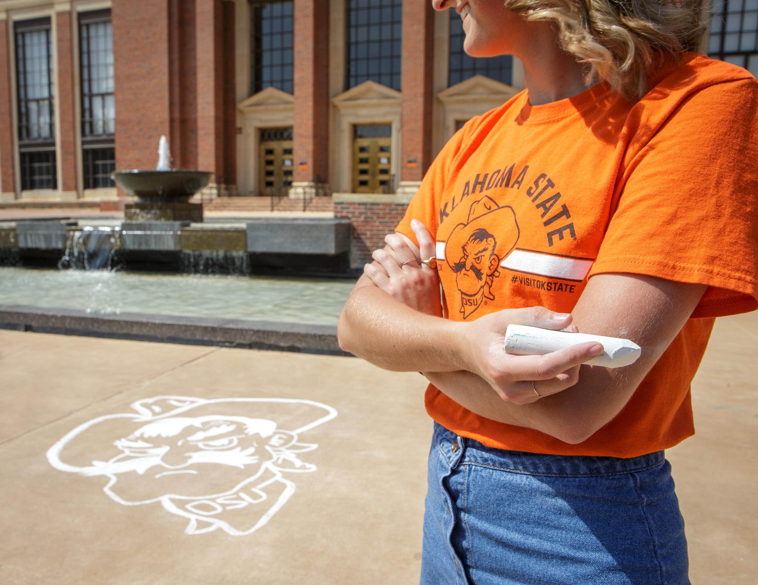 A young woman crosses her arms while holding a large piece of sidewalk chalk with a chalk drawing of Pistol Pete's face on the concrete behind her in front of Edmon Low Library and the fountain.