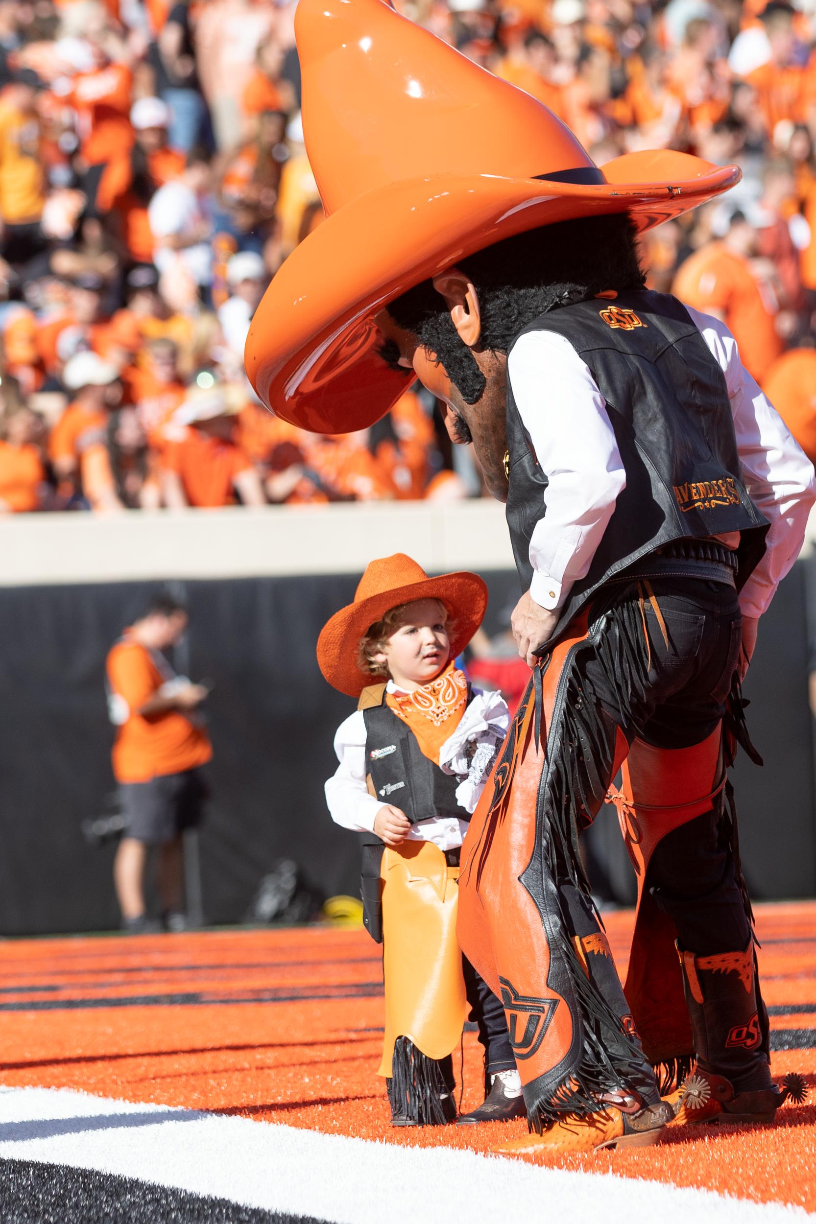 Pistol Pete stands on the field at Boone Pickens Stadium in the endzone while looking down at a young child who is dressed like him.
