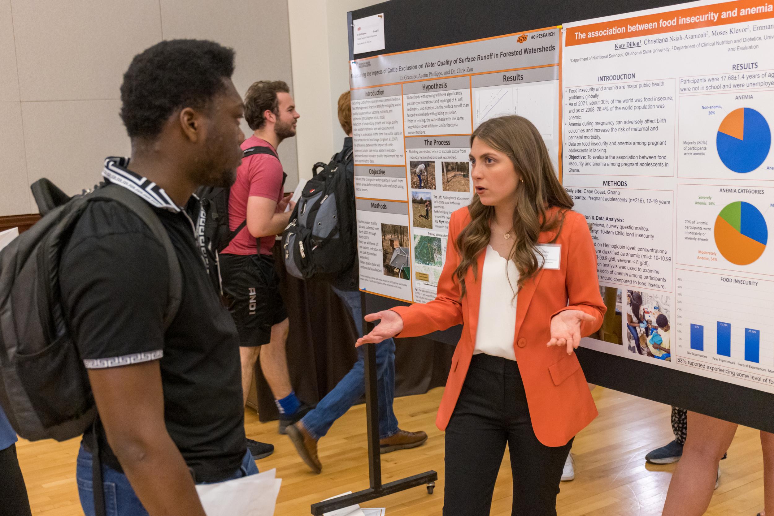 A white female student wearing an orange blazer explains her research poster to symposium attendees.
