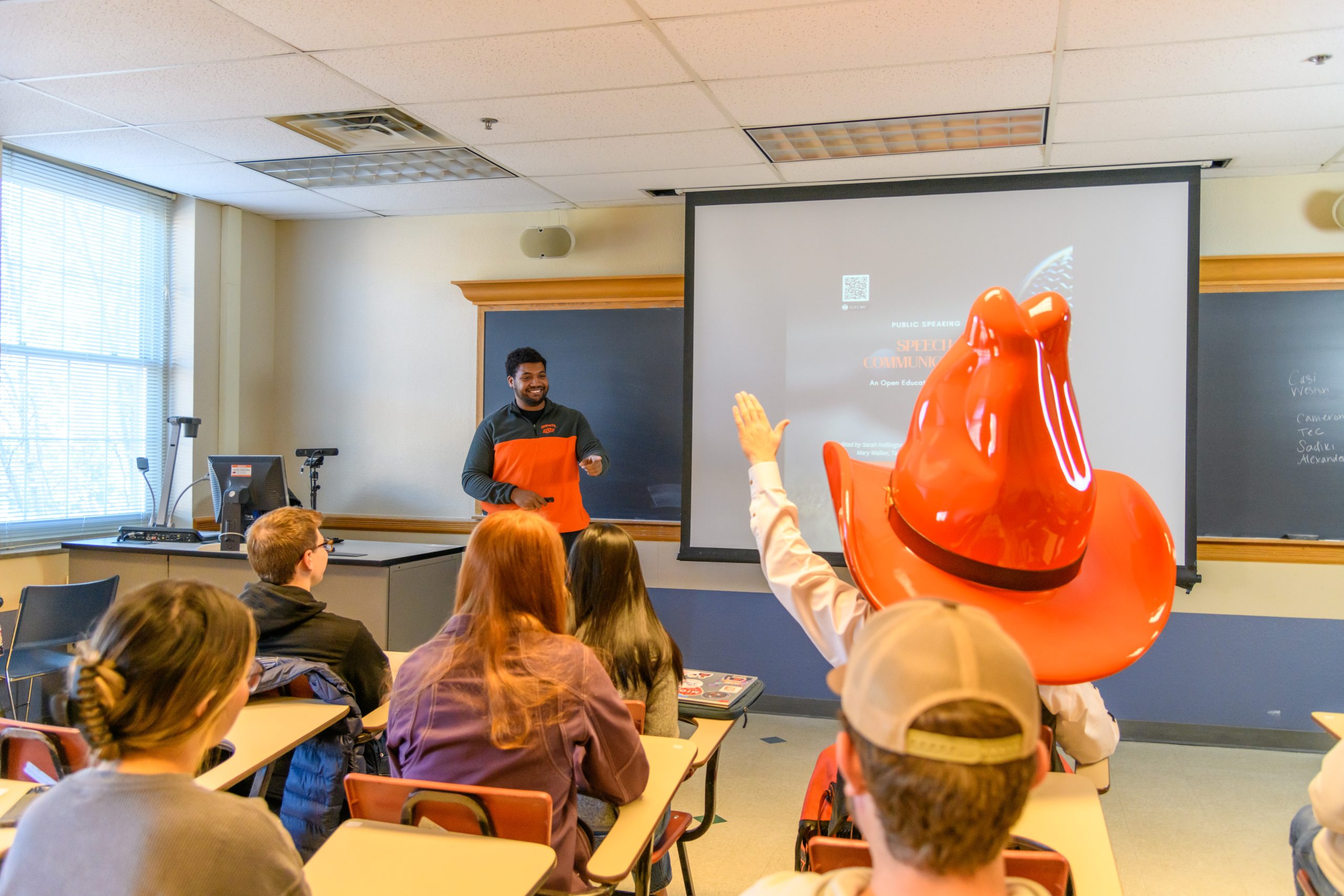 Pistol Pete sits in a desk in a classroom, while raising his hand to ask the speaker a question.