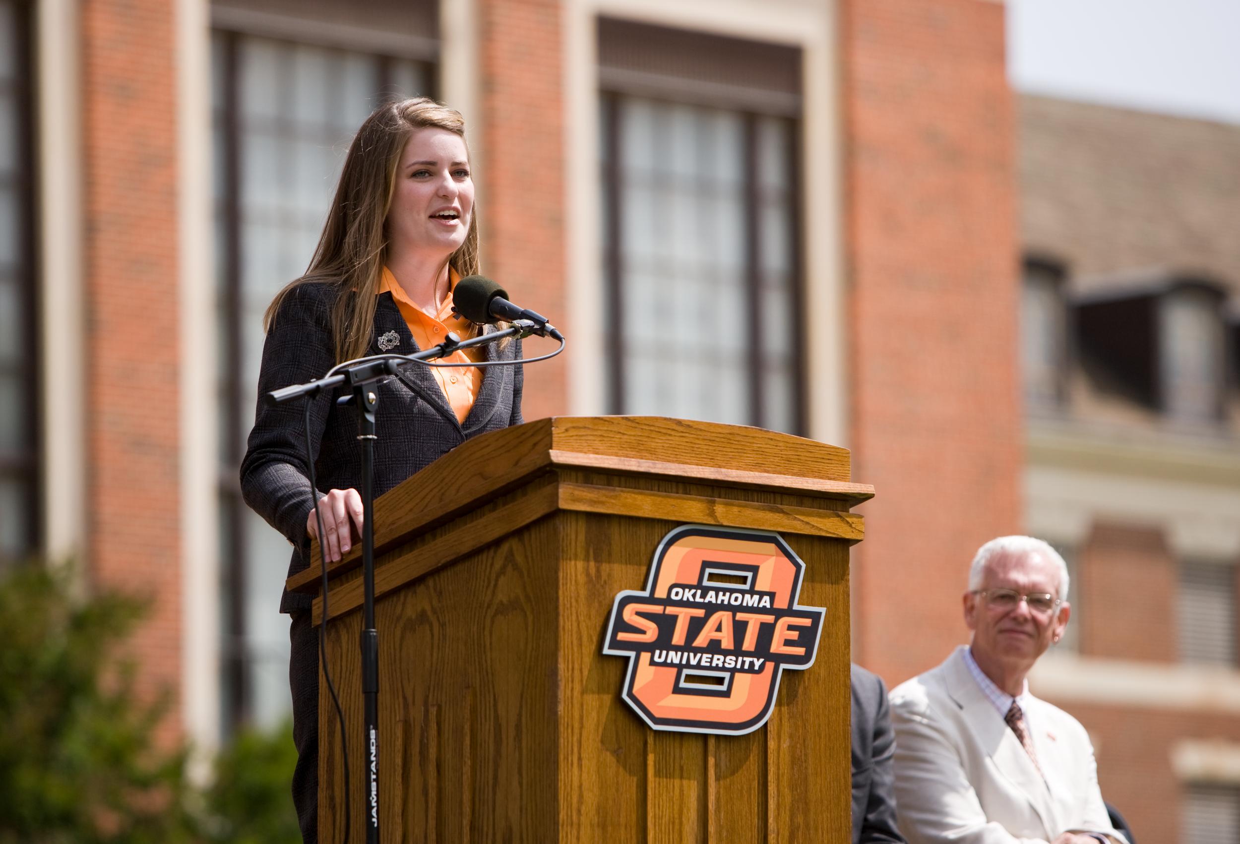A young woman speaks at a lectern with an Oklahoma State logo on the front.