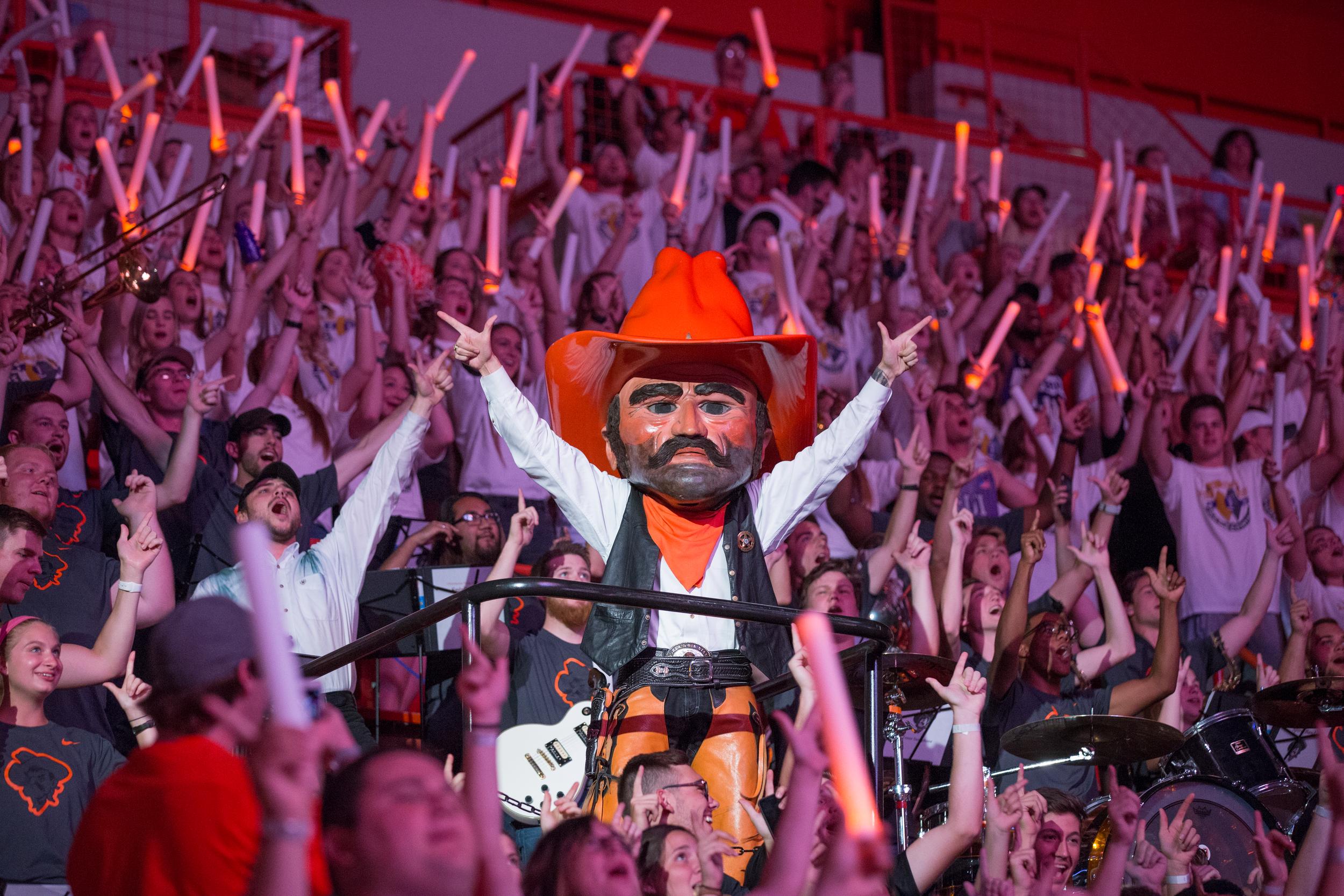 Pistol Pete stands on a platform at OSU's homecoming & hoops event, surrounded by students holding orange glow sticks and showing the go pokes gesture.
