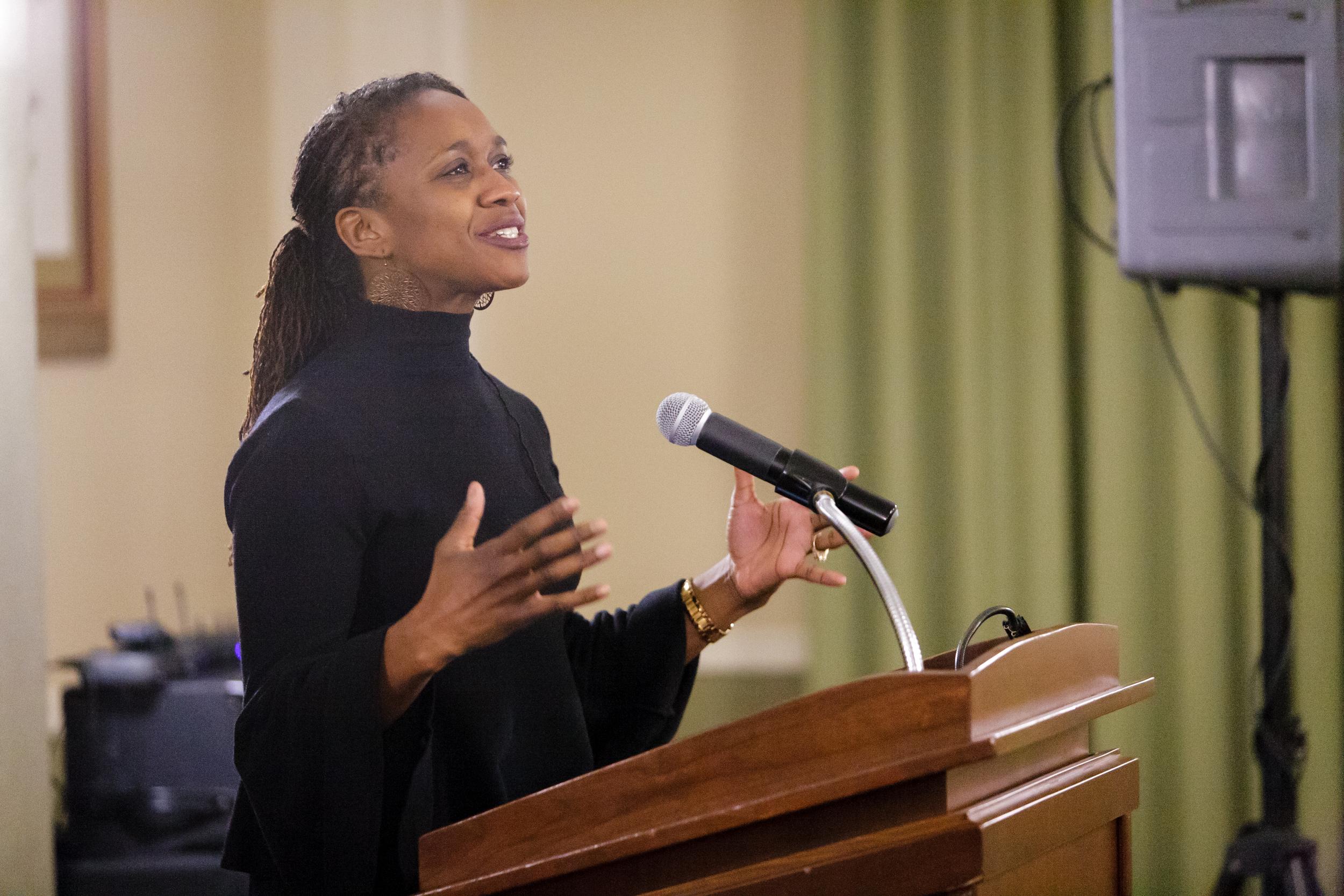 A black woman in a black sweater speaks behind a lectern while simultaneously using her hands to gesture effectively