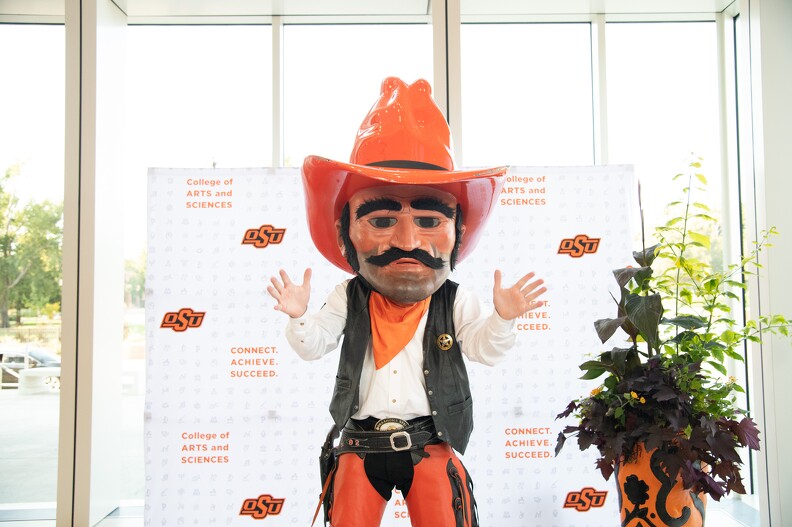 Pistol Pete stands in front of a backdrop and a plant, placing his hands straight out in front with his palms facing forward.