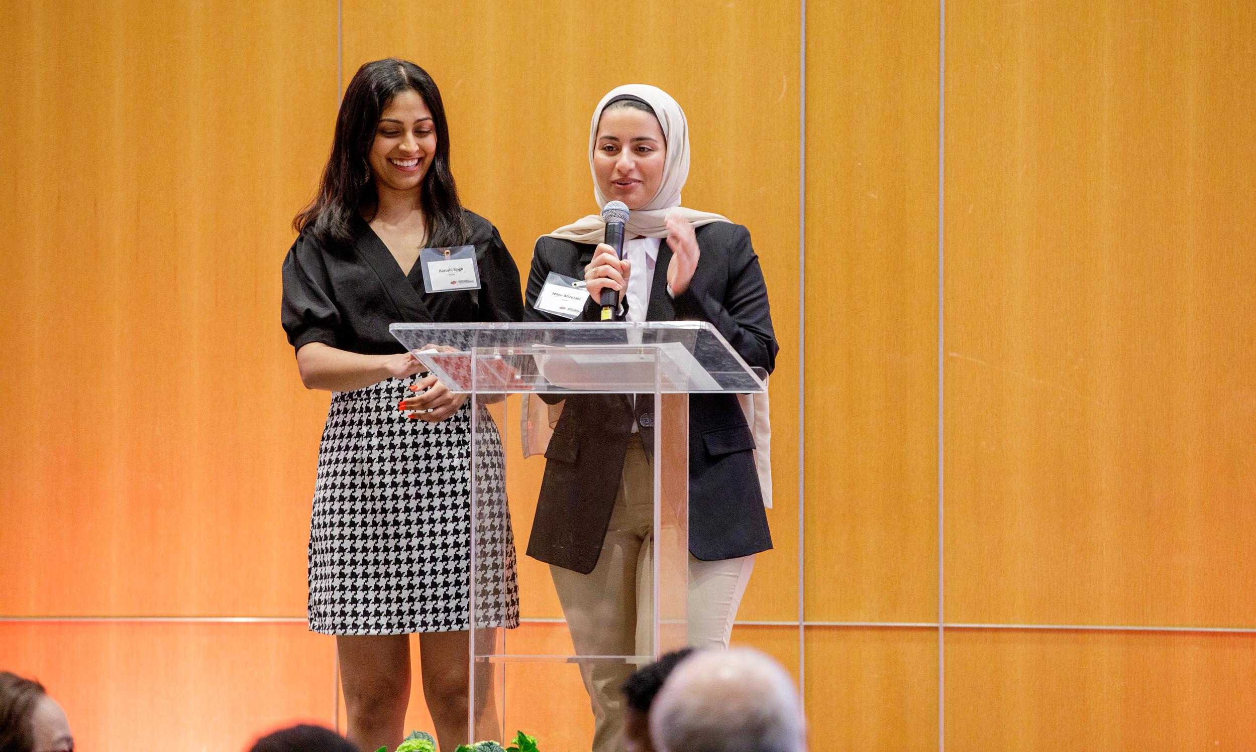 Two women stand at a clear lectern with a microphone to talk to an audience