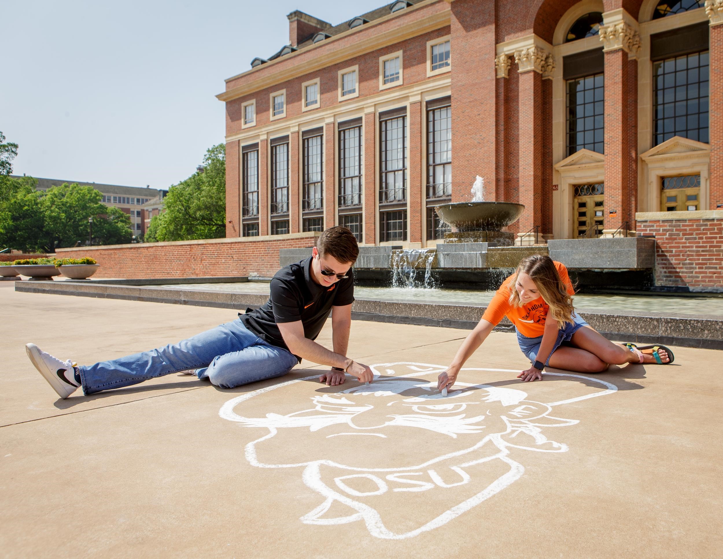 A man and a woman create a chalk drawing of Pistol Pete's head in front of the fountain at Edmon Low Library.