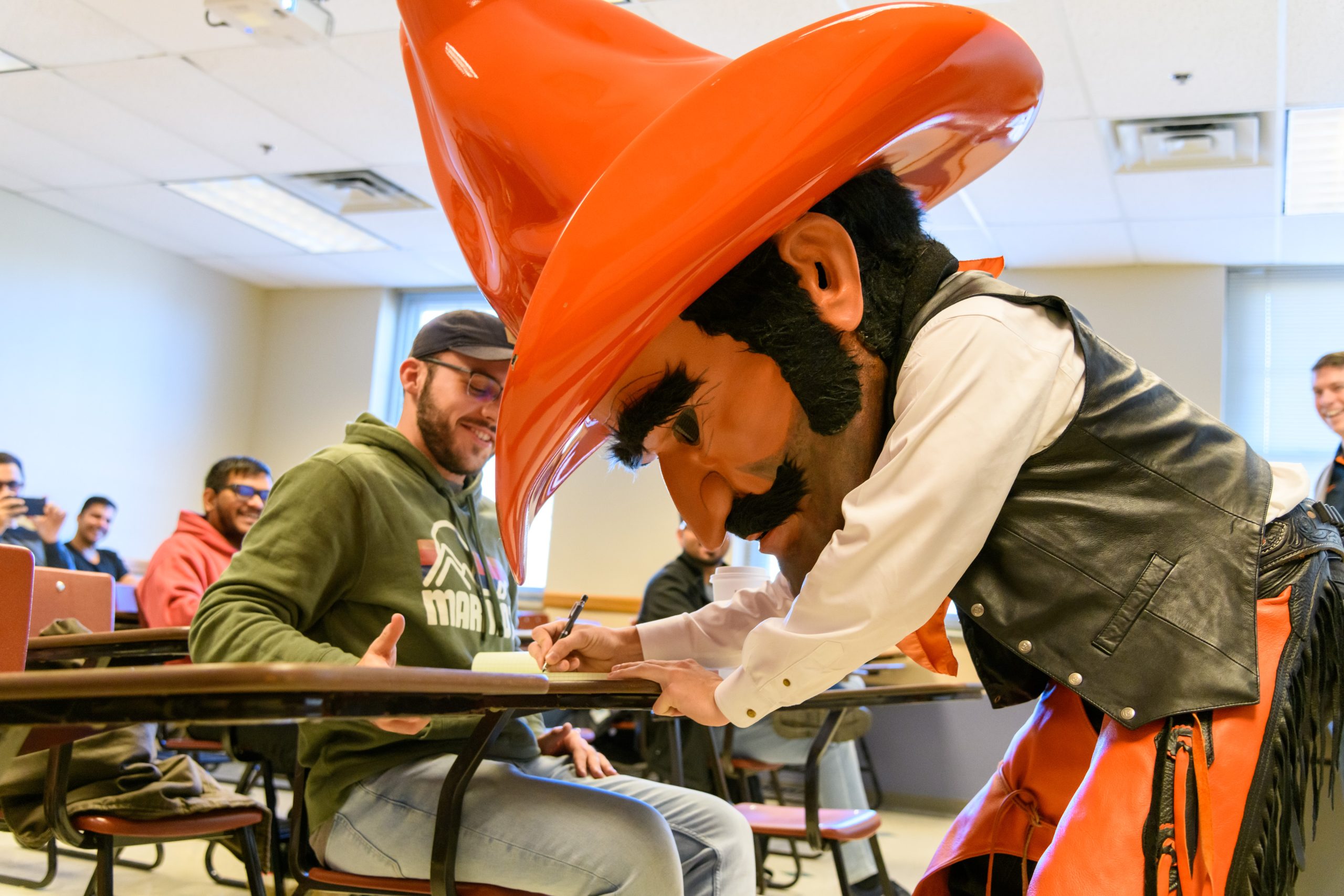 Pistol Pete writes on a paper on a desk in a classroom while standing up.