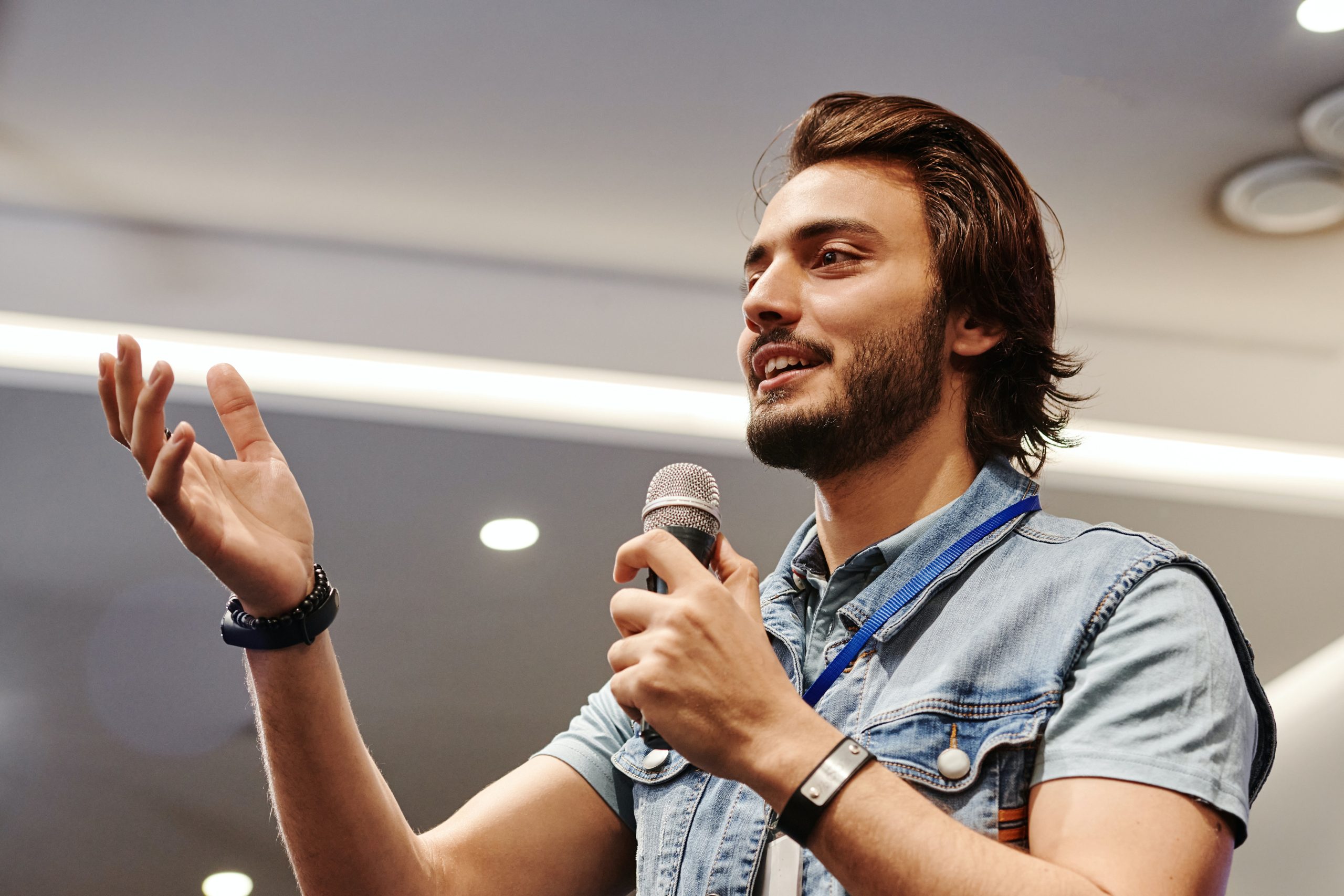 A young man holding a microphone smiles and gestures with his hand towards the audience.