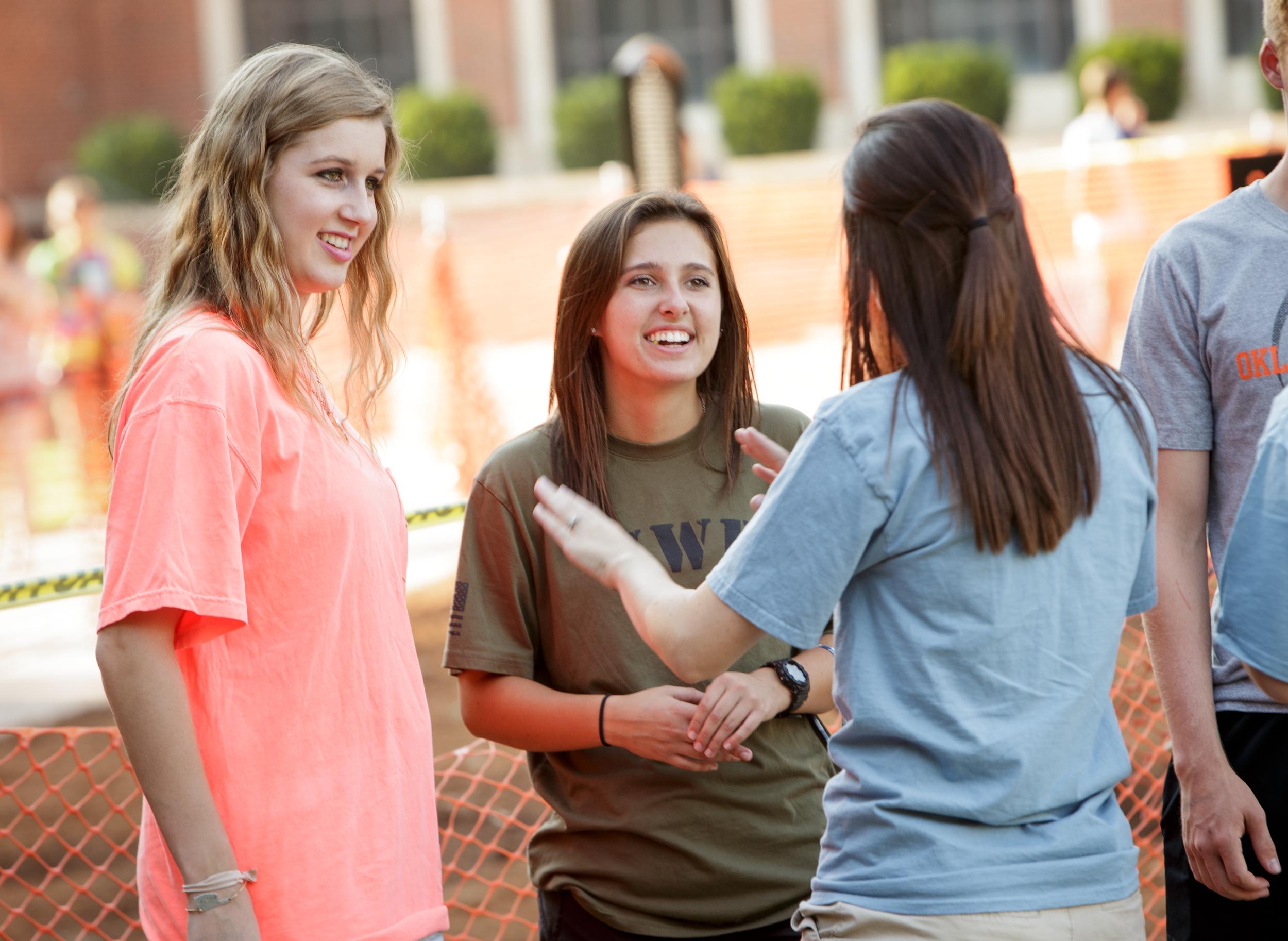 A young woman with long brown hair speaks to two other women in an outdoor campus setting