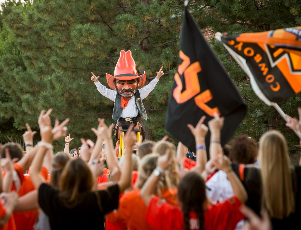 Pistol Pete and a crowd of OSU students hold their hands in the air displaying the gesture for "Pistols Firing/Go Pokes."