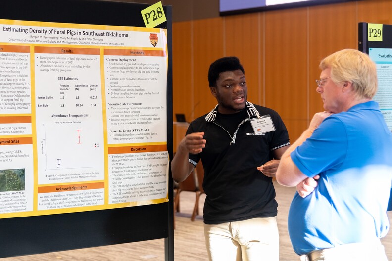 A man explains a poster presentation aid to another man at a research conference.