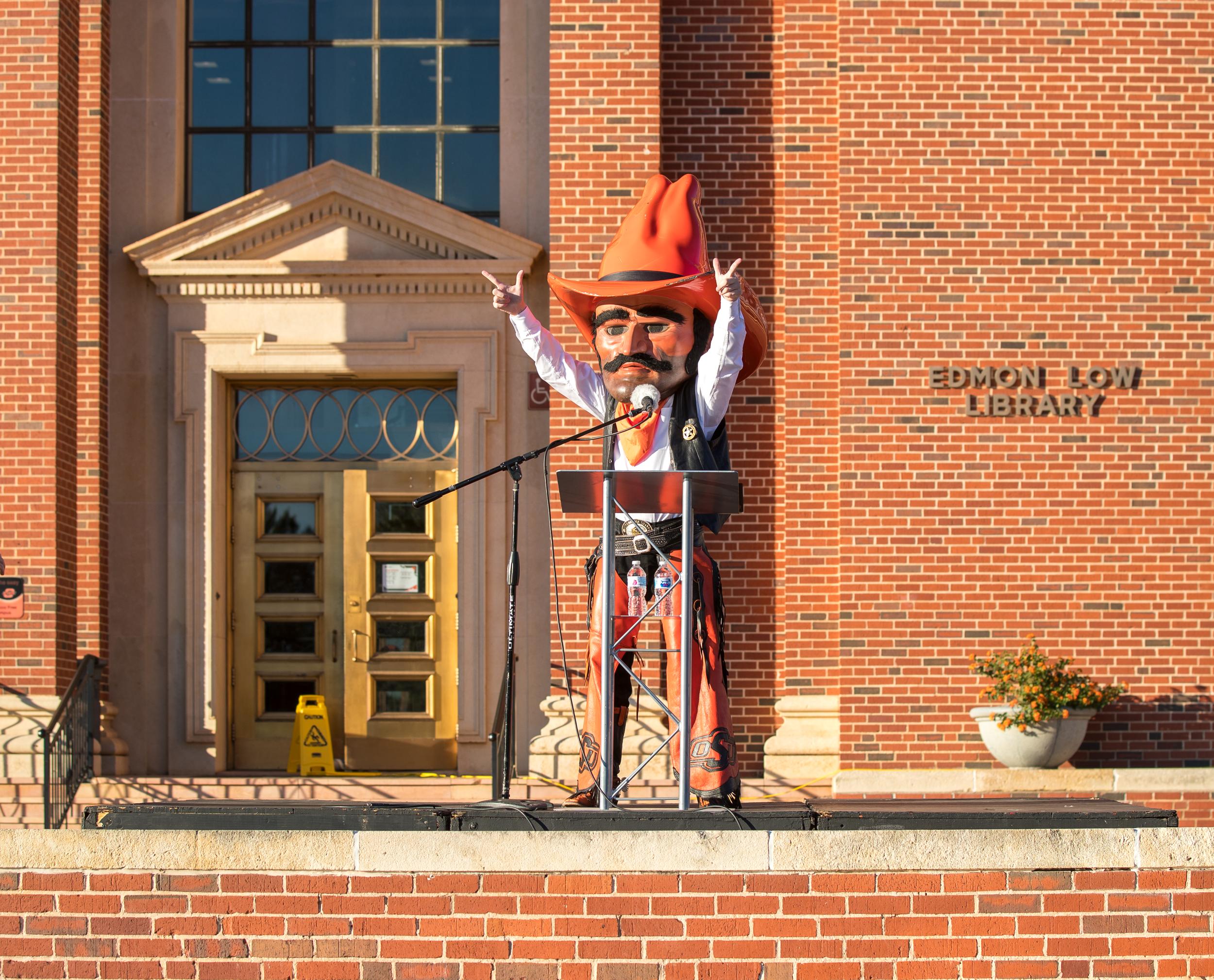 Pistol Pete stands at a lectern with a micophone in front of Edmon Low Library while proudly showing "Go Pokes" with both hands.