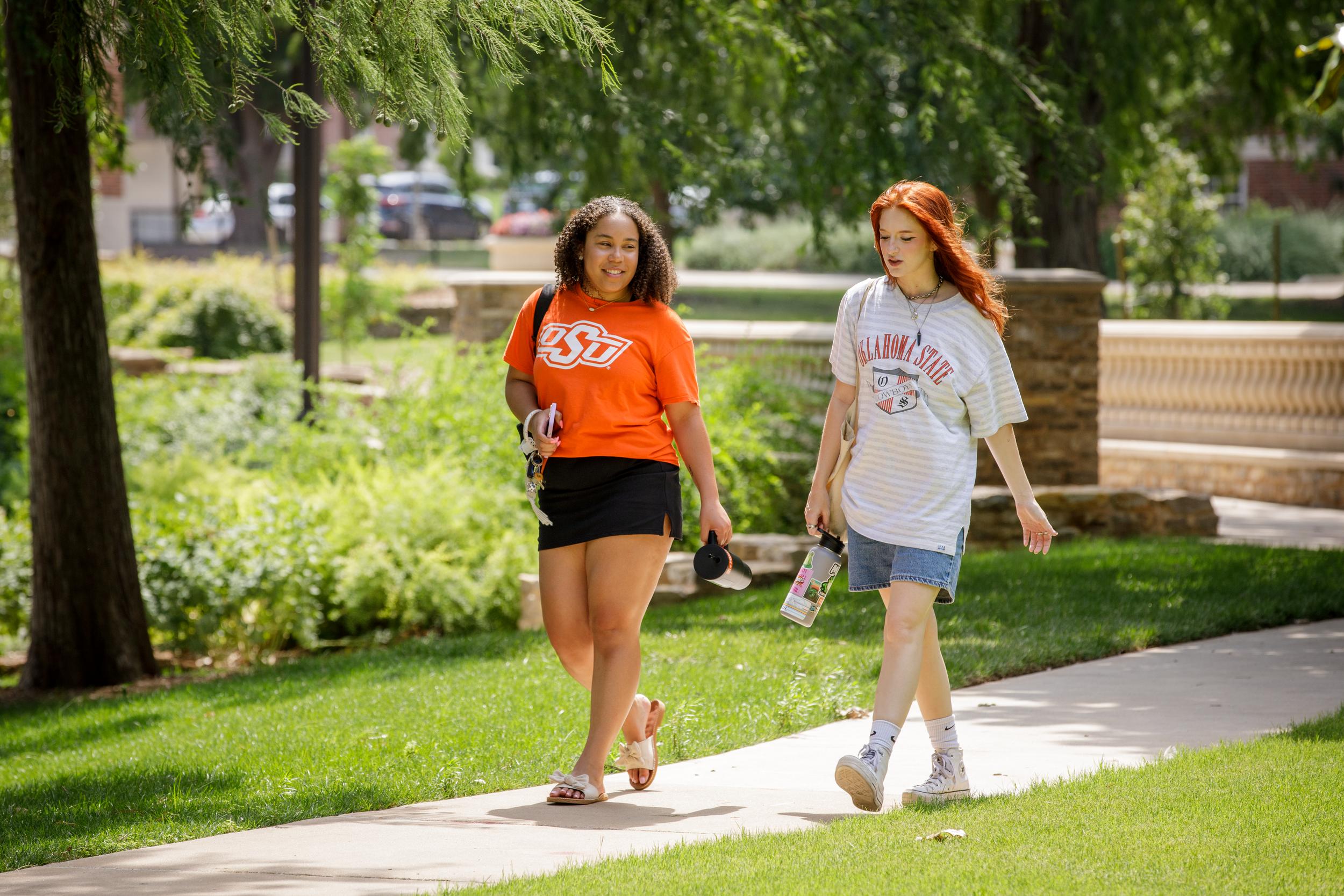 Two female college students are chatting while walking on a sidewalk.