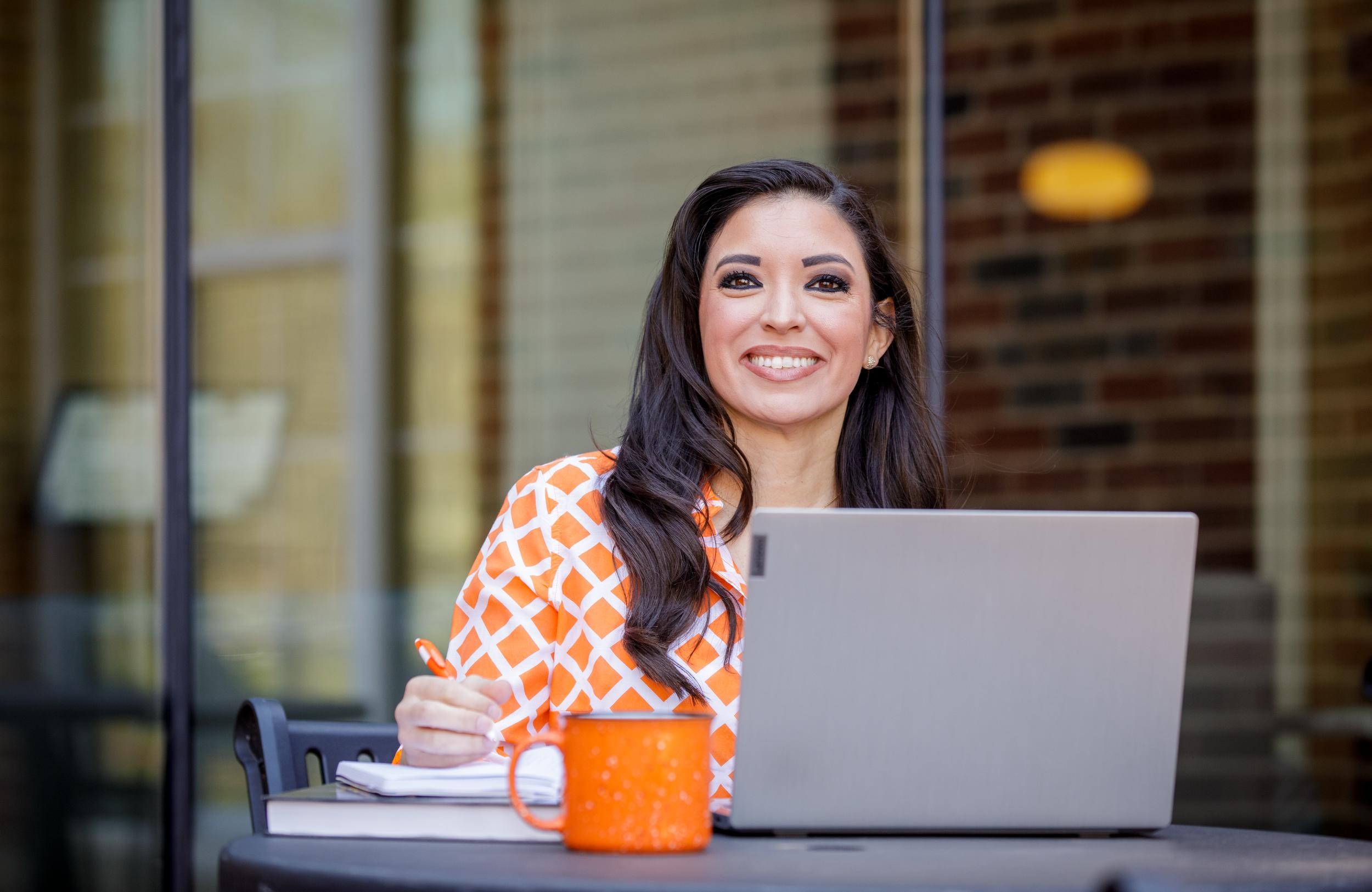 Young woman sitting in front of an open laptop smiles at the camera.