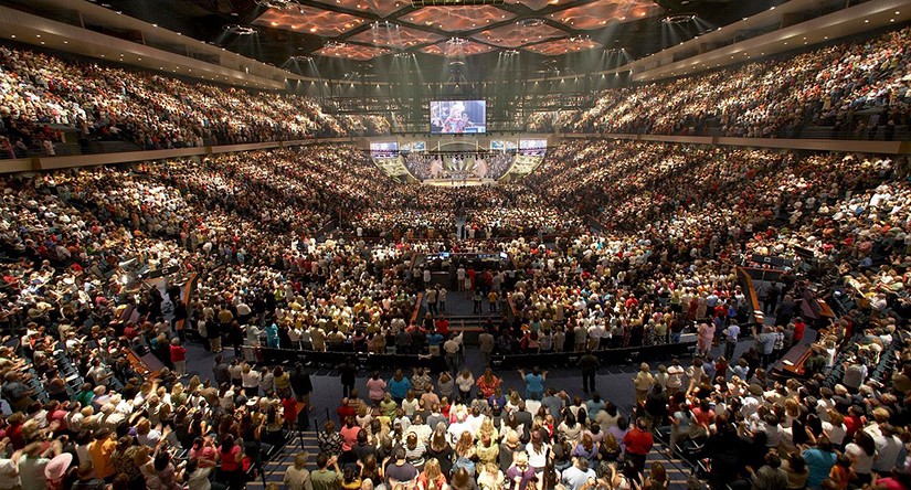 A church service at Lakewood Church in Fort Lauderdale, Florida. It looks similar to a large concert venue with thousands of people in seats, large video screens, and a stage in the center.