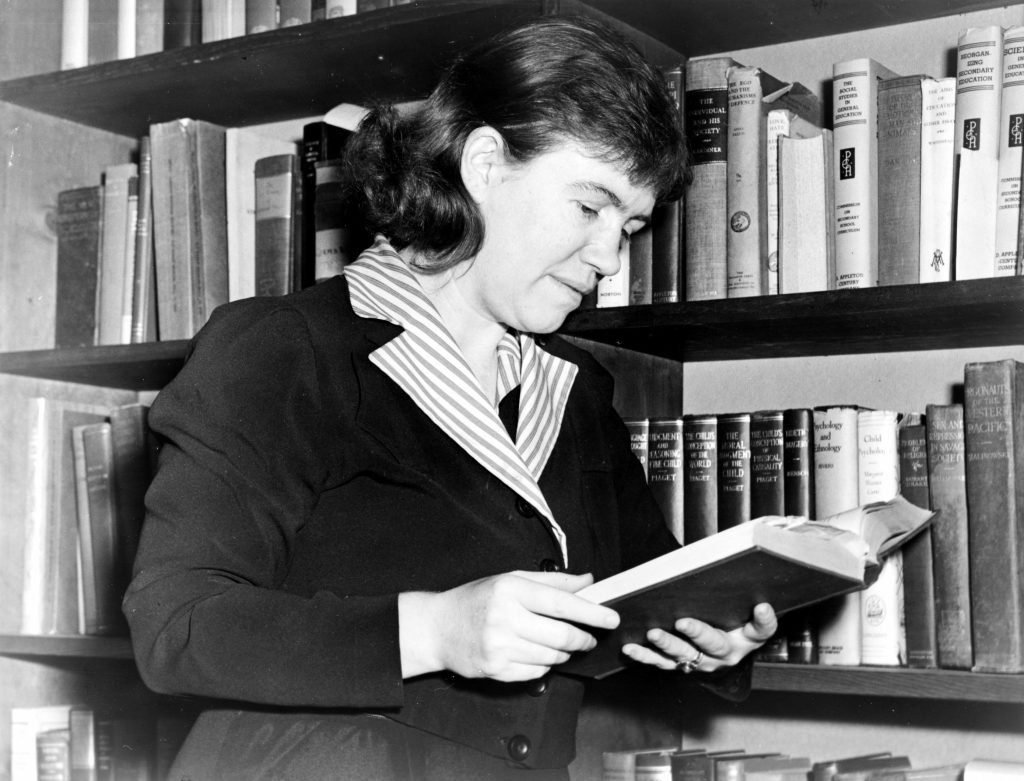 Margaret Mead reading a book while standing next to a shelf of books.