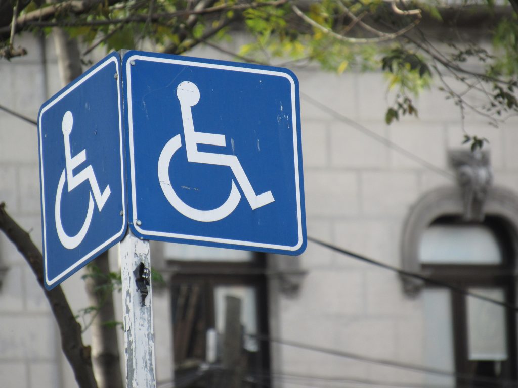 An image of a handicap parking sign in front of a tree and a gray stone building.