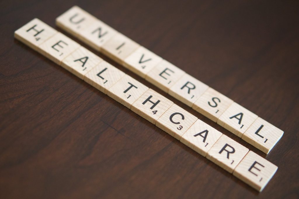 An image of tan wooden Scrabble game tiles, arranged to spell the words "Universal Healthcare" against a dark wood background.