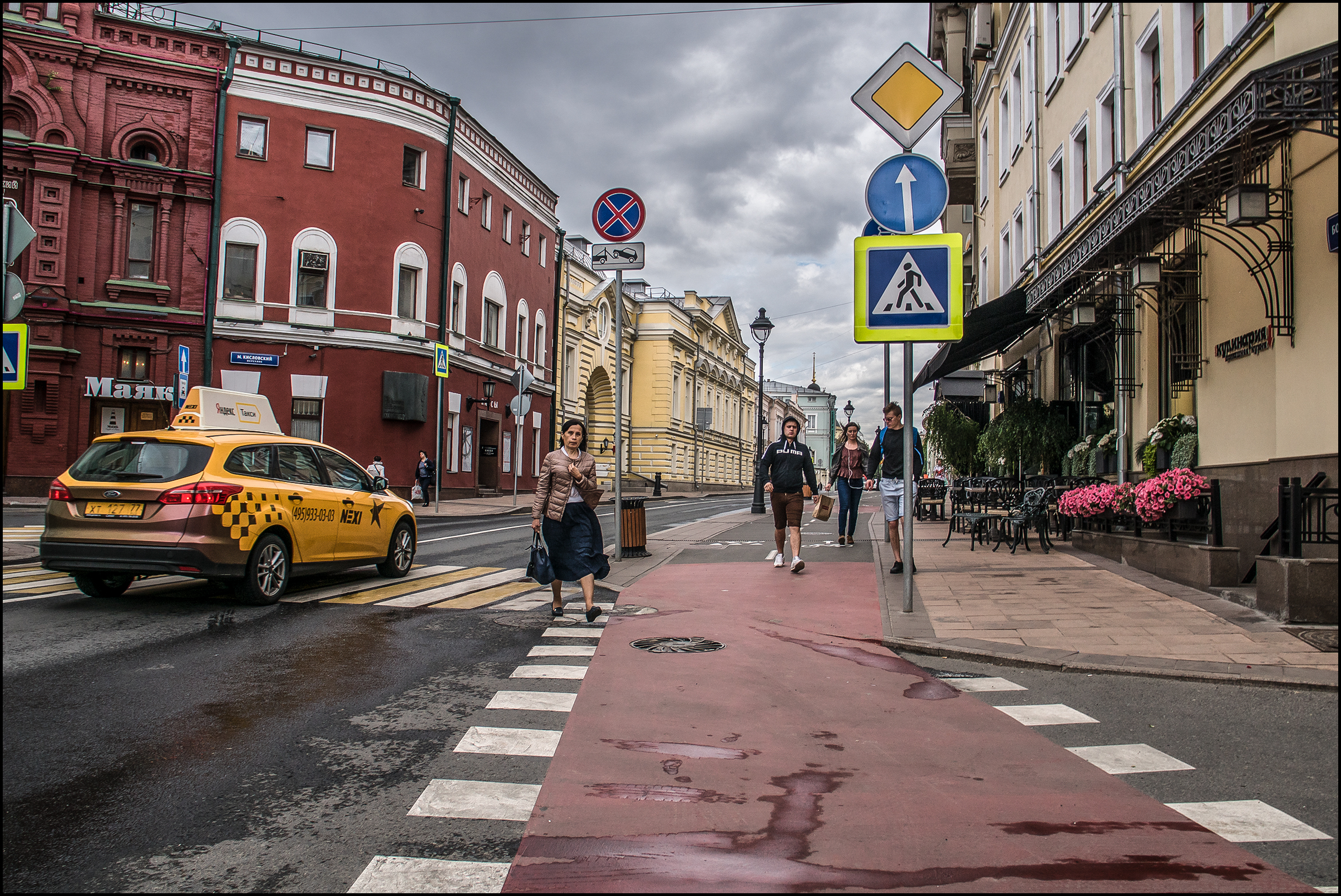 A photo of people walking along a street in Europe, with a yellow taxicab on the left and a sidewalk with tables and flowers on the right. There is a crosswalk sign in the foreground.