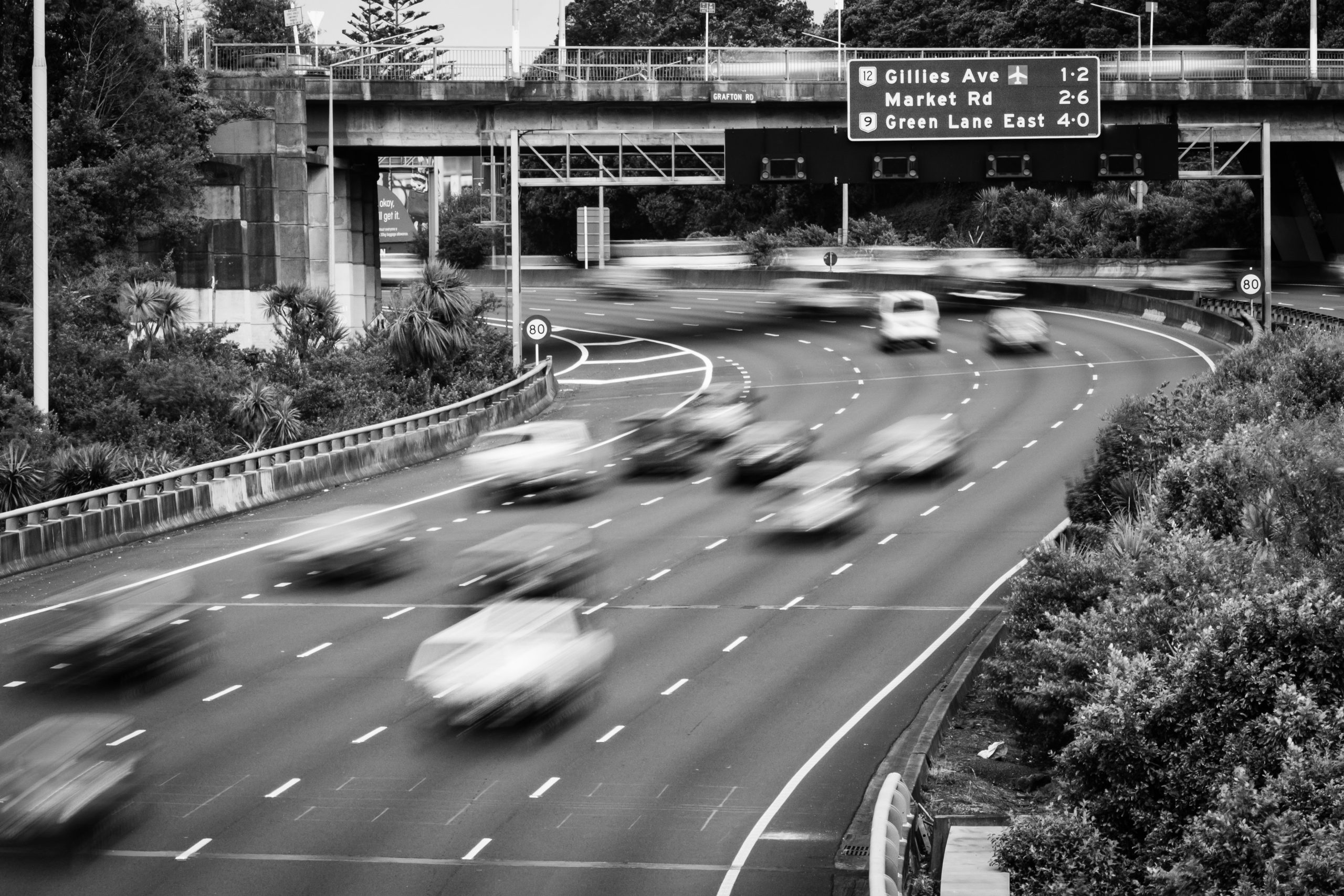 A black and white photo of blurry cars on a highway road. The road curves under an overpass with a sign that says "Gillies Ave 1-2. Market Road 2-6. Green Lane East 4-0."