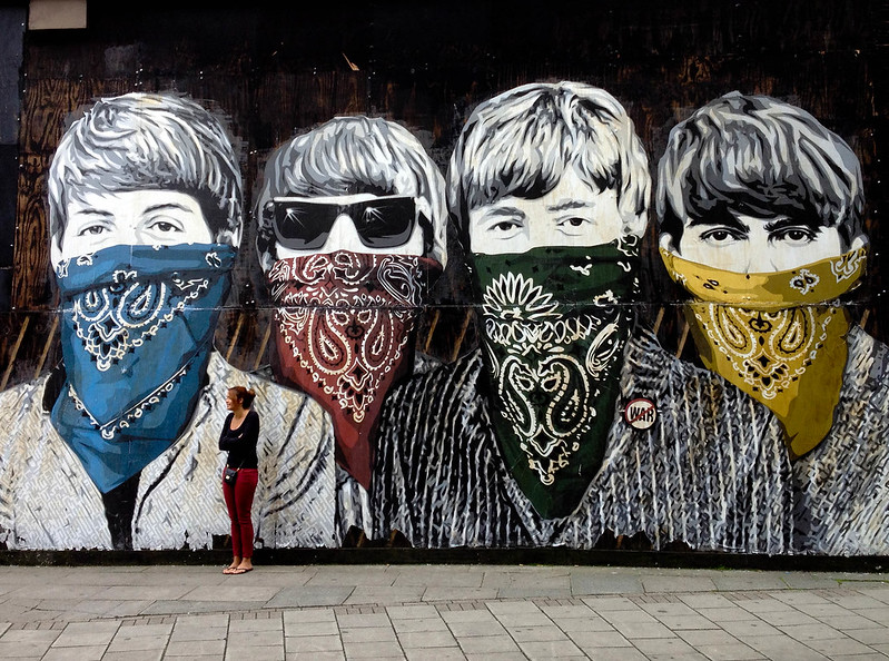 A girl standing in front of a wall with a large artwork of The Beatles, with each member of the band wearing a colored handkerchief.