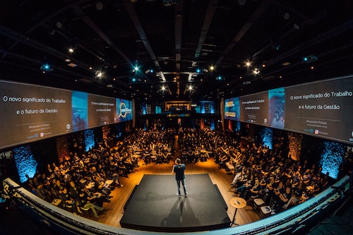 A room with roughly 200 people sitting and watching a speaker on stage.