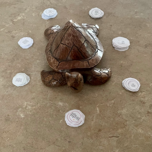 A carved wooden turtle sits on a stone surface surrounded by seven small stacks of round white paper pieces arranged in a circle.