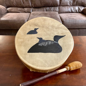 A hand drum with a natural tan surface features a black painted design of three loons, one large, one small one on its back, and one smaller above them. A drumstick with a padded sueded head rests in front of the drum on a wooden table, with a brown leather couch in the background.